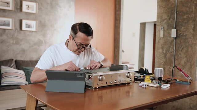 Mature Asian man disassembling audio equipment and self-learning, searching for manual guide to fix circuit board in tablet, home appliance DIY, electronics component hobby, handyman skill workshop.
