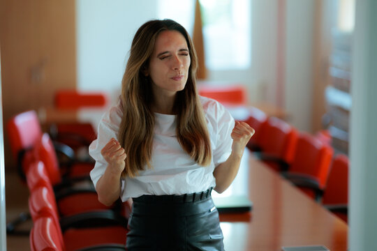 Angry Businesswoman Clenching Fists In Frustration Standing Inside Empty Conference Room. Annoyed woman in a white blouse stands celebrating or expressing rage