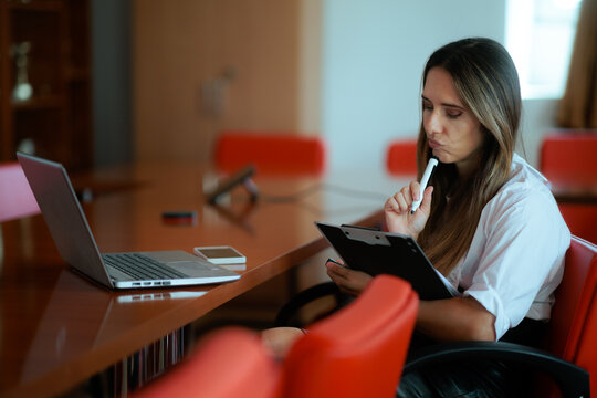 Pensive Businesswoman Holding Clipboard And Pen Sitting At Conference Room Desk. Thoughtful woman looks down while holding a clipboard next to a laptop