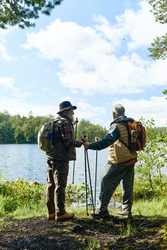 Vertical image of Caucasian senior men hiking with trekking poles near lake. Active retirement concept suitable for healthcare campaigns and tourism advertising