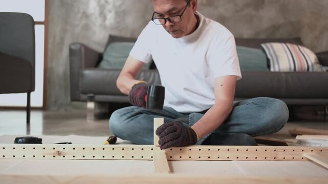 Asian mature man self-assembling flat-pack furniture with DIY tools, nailing joint parts with rubber mallet, home improvement, craft installation, hobby wooden workshop, handyman active lifestyle.