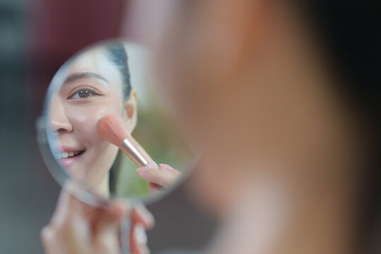 Asian woman applying blush with makeup brush in mirror for cosmetics beauty routine