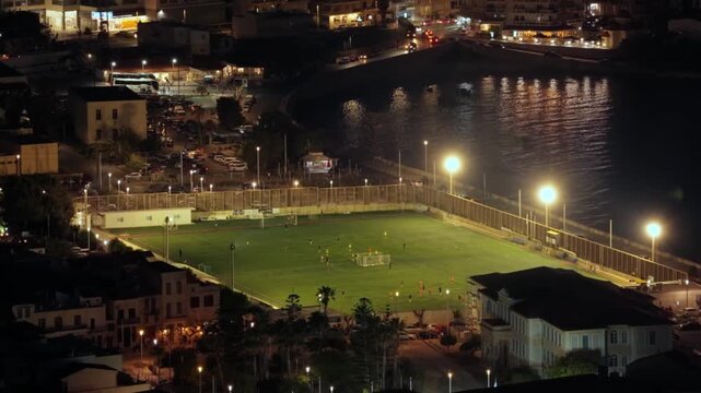 Aerial Night View of Football Stadium Near Harbor With Players on Green Pitch in Greece