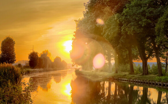 sunset over canal with golden light reflecting. Trees line calm bank while warm sun creates lens flare and mirror-like water,