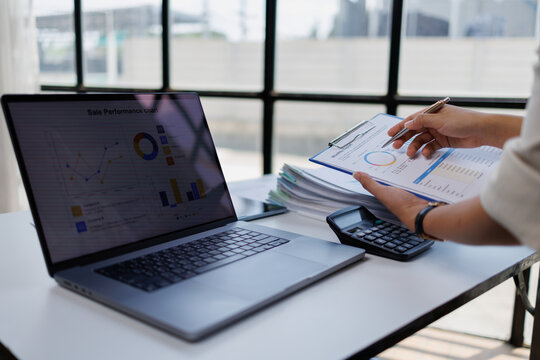 Professional hands reviewing financial reports and sales charts on a clipboard, comparing data with a laptop screen displaying analytics, next to a calculator