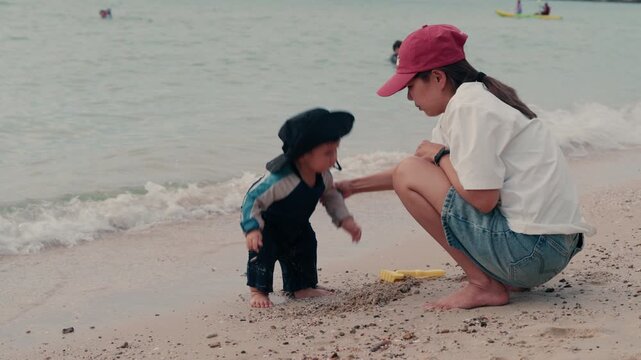 Happy mother and adorable baby boy sitting on sandy beach together during summer vacation