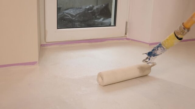 A worker uses a long-handled paint roller to apply an even coat of primer on a floor near a window, masking tape protecting the walls