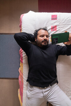 A top-down view of a man lying on a bed in a bedroom, checking his cell phone