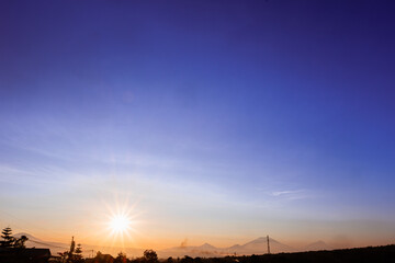 Obraz premium A stunning sunrise landscape with a bright sunburst effect on the eastern horizon, showing the silhouette of the countryside, trees