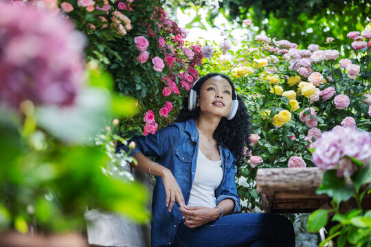 A woman wearing headphones sits among colorful blooming roses and lush greenery, looking upward in a sunlit garden. The peaceful scene captures personal downtime, outdoor relaxation