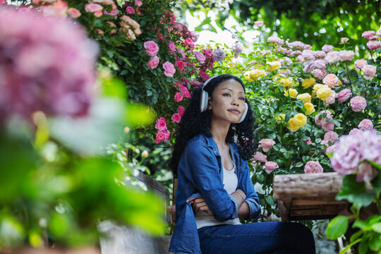 A woman wearing headphones sits among colorful blooming roses and lush greenery, looking upward in a sunlit garden. The peaceful scene captures personal downtime, outdoor relaxation