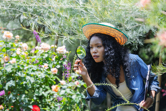A woman wearing a straw hat closely examines garden flowers with a magnifying glass. Her focused expression highlights attention to detail, thoughtful plant care, and curiosity about nature.