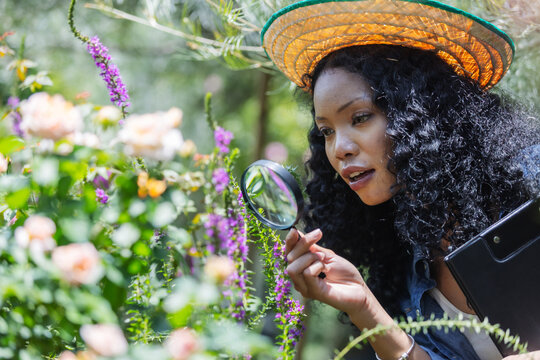 A woman wearing a straw hat closely examines garden flowers with a magnifying glass. Her focused expression highlights attention to detail, thoughtful plant care, and curiosity about nature.