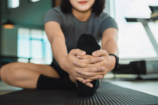 Young woman stretching leg forward while sitting on exercise mat indoors in modern fitness studio during workout session