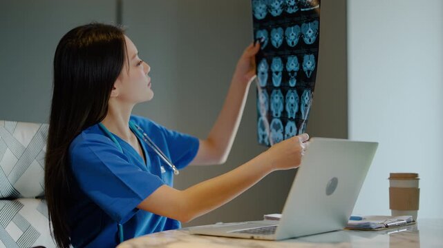 Young Asian female doctor analyzing brain x-ray CT scan film while typing on laptop at desk in hospital office. Professional medical worker reviewing patient radiograph and health data online.