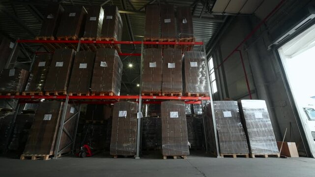 Tall warehouse racking filled with palletized cardboard boxes wrapped in clear plastic beside bright loading doorway on concrete floor
