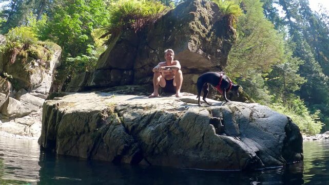 A man and his dog sit on a large boulder in Inter River Canyon in North Vancouver, British Columbia, Canada, surrounded by rocky terrain and forested natural scenery