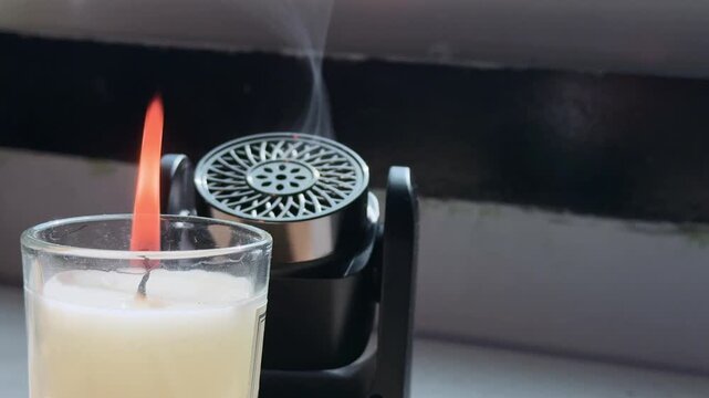 Close-up of a burning white candle in a glass jar next to a black aroma diffuser emitting a thin mist. The objects sit indoors on a windowsill illuminated by bright natural daylight.