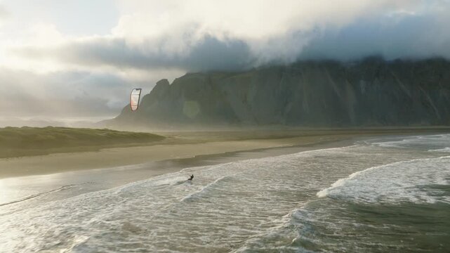 Kite surfer follows Atlantic shoreline beneath distinctive Vestrahorn mountain ridge, with dark coastal sand, breaking waves and low cloud covering upper slopes at Stokksnes, Southeast Iceland, aerial