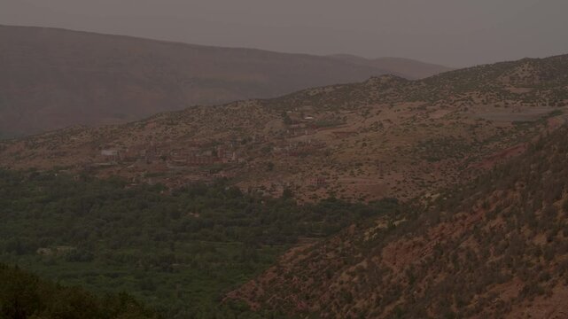A panoramic view captures a lush, forested valley floor rising into rolling arid foothills and distant Berber settlements under the warm, hazy mountain sky of Morocco&rsquo;s picturesque High Atlas range.