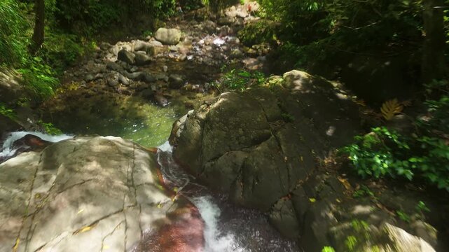 The camera flies low along a boulder filled stream above the Cascades de Didier, Martinique, clear water sliding between rocks before dropping away out of sight. Aerial flyover shot
