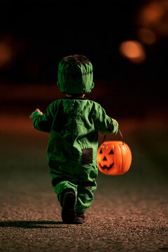 Frankenstein's monster on Halloween night as toddler walks away carrying orange pumpkin bucket while trick or treating