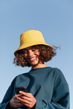 Smiling woman with curly hair wearing yellow bucket hat checking mobile phone outdoors for lifestyle banner and social media background design