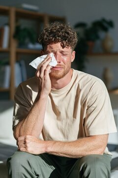 Upset man wiping tears with tissue while sitting on bed background for emotional wellness banner and health awareness design