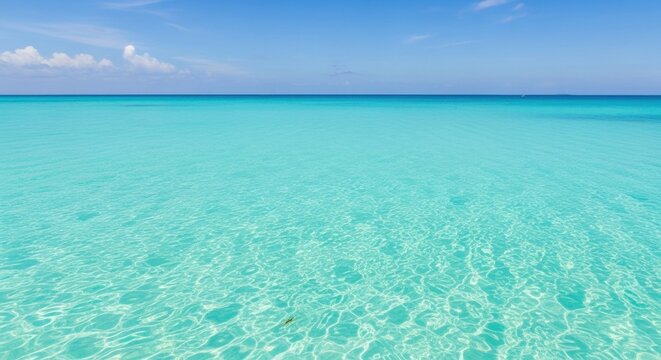 Clear turquoise ocean water with gentle ripples on the surface under a bright blue sky with soft white clouds, featuring a clean horizon line and transparent tropical sea.