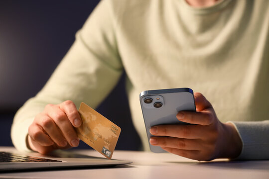 Young woman with credit card and mobile phone using laptop for online shopping at table in evening, closeup
