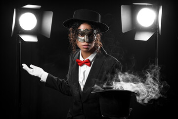 Young African-American magician in mask showing magic trick with hat on black background © Pixel-Shot