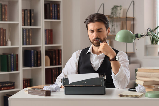 Young male writer with vintage typewriter, books, crumpled papers and smoking pipe working in office