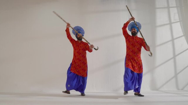 Two Indian Bhangra dancers are energetically performing with sticks in a studio setting