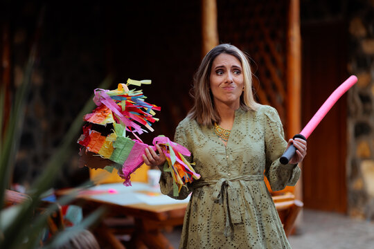 Funny Woman Holding Colorful Pinata And Stick Looking Confused At Party. Playful lady shrugs while holding a pinata toy and pink stick outdoors