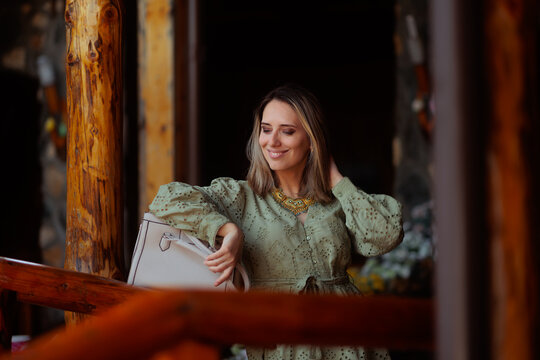 Smiling Elegant Blonde Woman Wearing Stylish Green Dress Standing On Veranda. Happy blonde with her handbag leaning against a wooden porch railing
