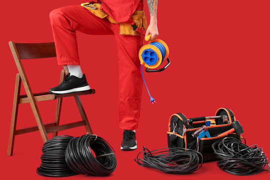 Male electrician with extension cable reel, wires and tool bag on red background