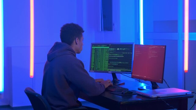 A young man with dark hair wearing a hooded sweatshirt types code on a keyboard at a desk with dual monitors in a neon-lit indoor workspace at nighttime.