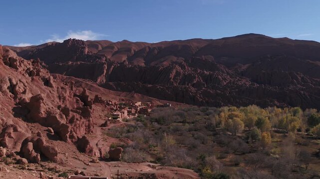 Panoramic aerial view showcasing a traditional clay-brick village nestled beside lush green oasis fields at the base of red rocky hills in Tinghir, Morocco.