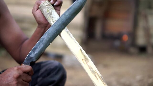 Close-up shot of a rural man's hand peeling and whittling wood using a sharp machete outdoors.