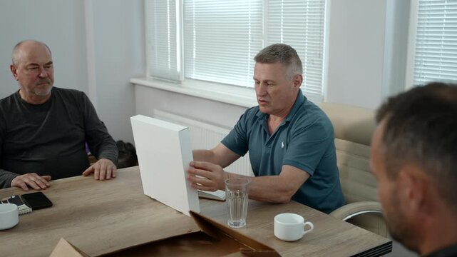 Older men continue focused office meeting with blank folder presentation, foreground observer blurred at right, and natural light filling clean modern room with blinds