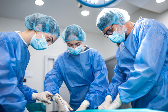 Expert medical team performing a surgical operation in a hospital. Skilled doctors and female surgeons in sterile blue gowns and caps concentrated on conducting a precise clinical procedure together.