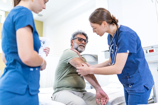 Pain in the arm. Arm pain, senior man with body-muscles problem, healthcare and medicine concept. Female doctor examining senior patient sitting on bed and holding painful arm in hospital room.