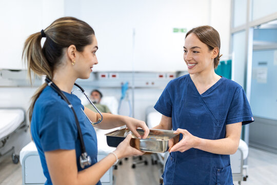 Two female medical doctors collaborating in a modern hospital ward. Professional female surgeons wearing blue scrubs discussing treatment while holding a metal medical tray near patient bed.