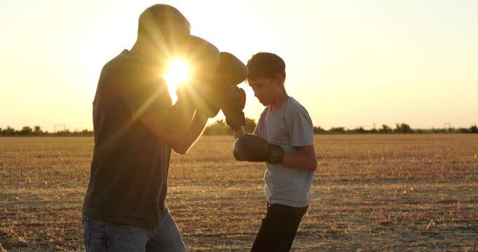 Father trains son in boxing drills with gloves across rural field. Parent and child practice punches with focus at sunset. Family fitness, mentorship and teamwork create sports footage.