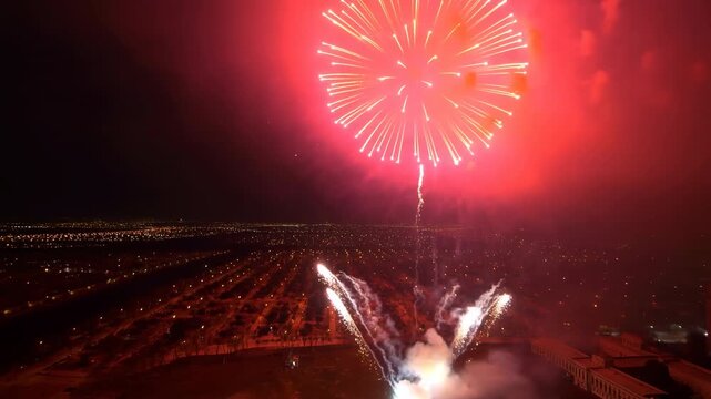 Colorful fireworks rise and burst above a broad nighttime landscape of lights, changing from golden trails to green and red spheres while smoke gathers around launch area