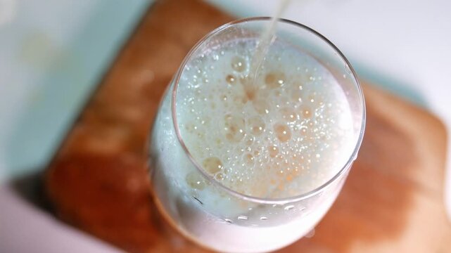 POV (zoom in): someone pouring a glass of beer on the table. Bubbles burst in the dense foam of the beer. A glass of draft beer on a table. A bar table. POV: drinking beer. A cold beverage on a table.
