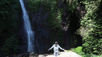 A person with outstretched arms stands on rocks in front of a majestic waterfall surrounded by dense green foliage under bright sunlight, enjoying the natural beauty. © Luthfia
