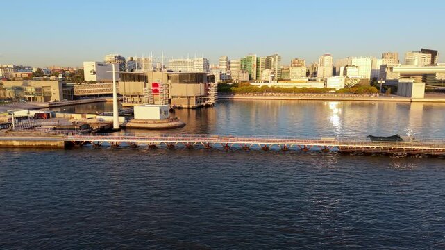 Cityscape by river at sunrise, calm waters reflect buildings. Long pier stretches into water, boats docked nearby. Modern skyline visible in distance. Peaceful, serene mood ideal for travel, urban