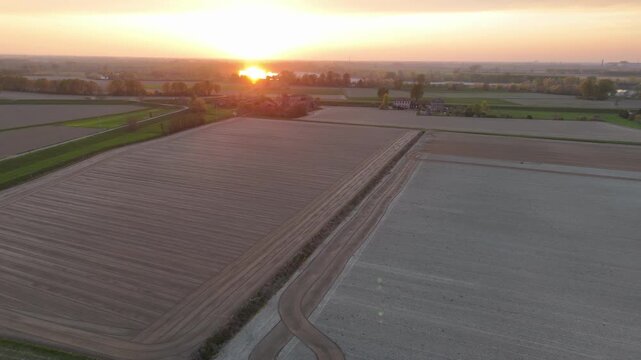 Freshly prepared Po Valley fields display parallel cultivation rows, curved farm tracks, drainage corridors, green parcels and distant farm buildings beneath warm sunset light, aerial shot.