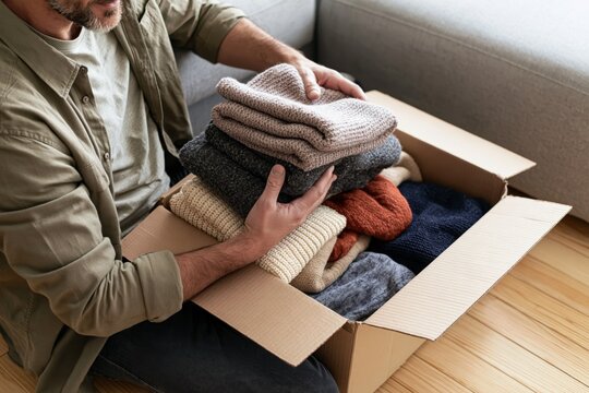 Man packing folded clothes into cardboard box for charity donation at home, concept of giving used clothing, decluttering, sustainable reuse and community support